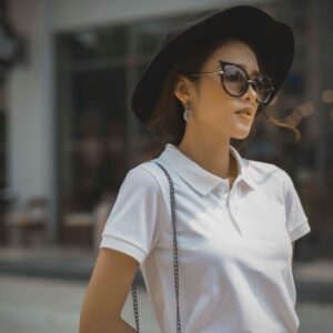 Fashionable woman in a white polo shirt, sunglasses, and black hat outdoors, exuding style and confidence.